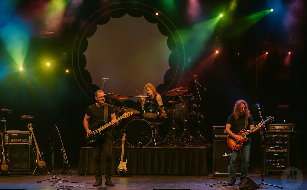 A band performs on stage, with a bassist in the foreground, a guitarist to the right, and a drummer in the background.