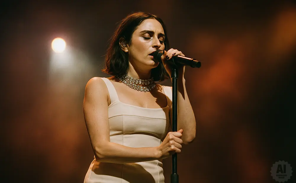 Singer performs on stage with microphone, wearing a dress and jeweled necklace, bathed in stage lights.