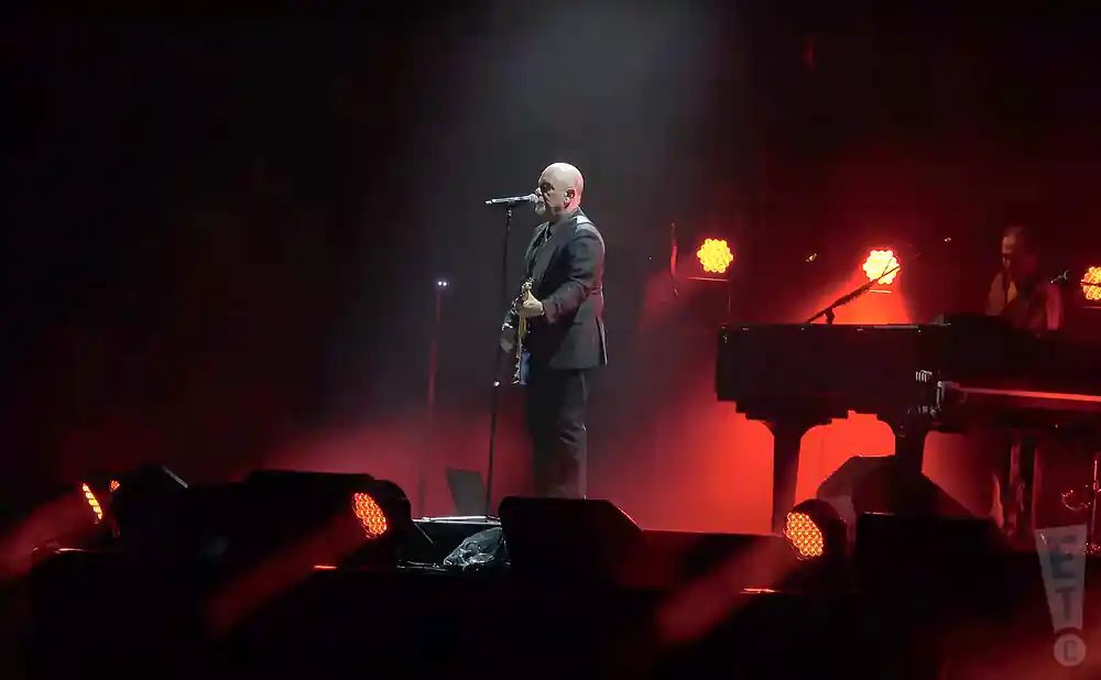billy joel on stage staring into the crowd in front of red and yellow lights