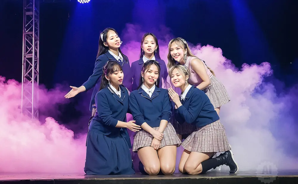 Six young women in school-like uniforms pose on a stage with purple lights and pink smoke.
