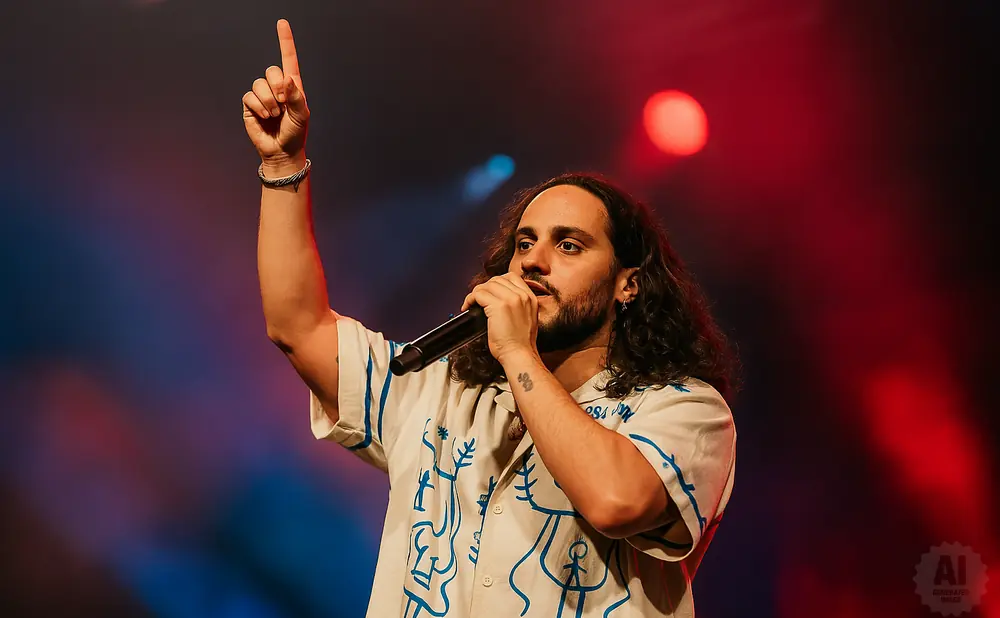 A male singer with long dark curly hair and a beard, wearing a light-colored patterned shirt, holds a microphone and points his finger up.