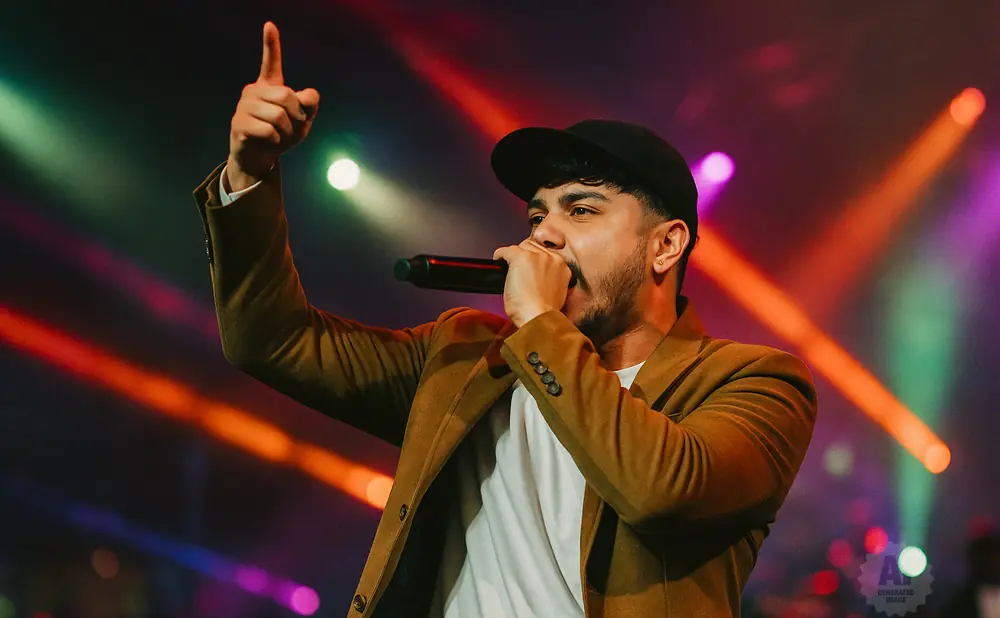 A man in a brown jacket and black cap sings into a microphone, pointing his finger upwards.