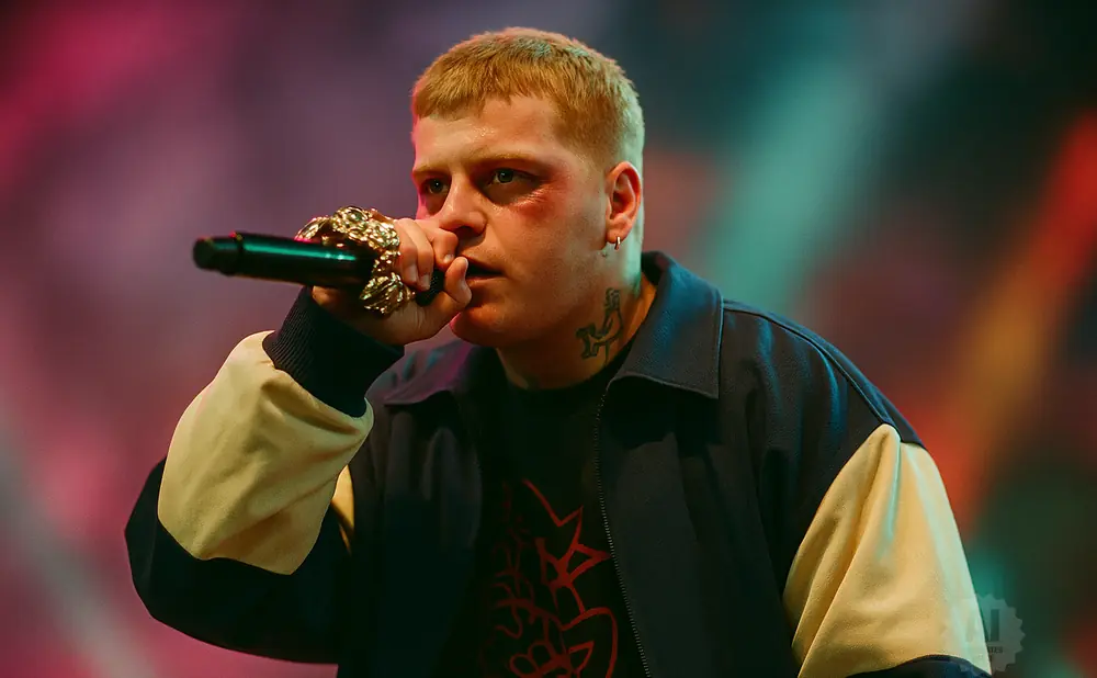 Rapper Yung Lean performs on stage, holding a microphone and wearing a blue jacket with yellow sleeves.