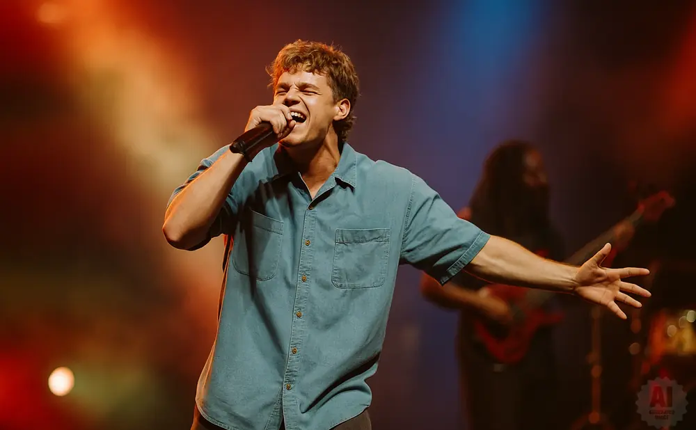 A male singer on stage, holding a microphone and singing passionately, with a blurred figure of a guitarist behind him.