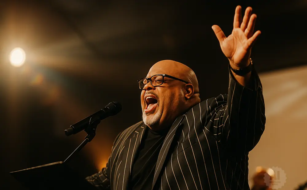 A Black man in a pinstripe suit sings passionately into a microphone, with one arm raised.