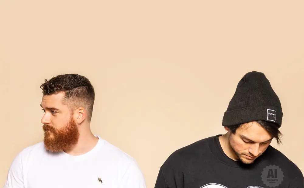 Two men against a beige background. The man on the left wears a white t-shirt and has a beard. The man on the right wears a black beanie and sweatshirt …