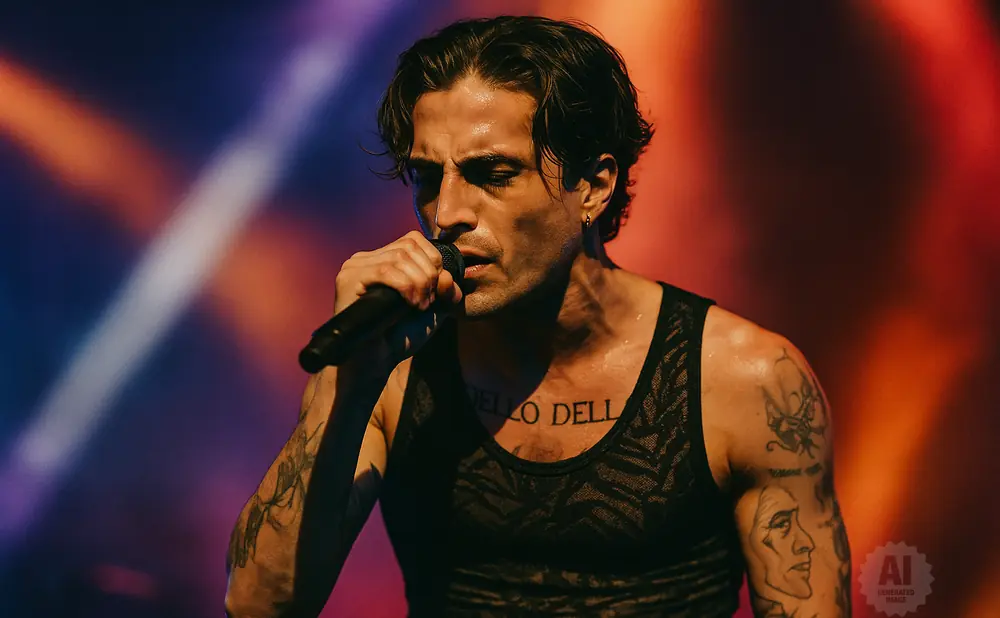 A male singer with tattoos performs on stage, holding a microphone and wearing a black tank top.