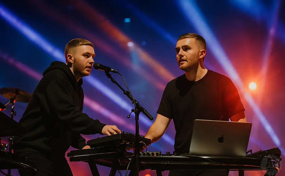 Two men perform music on stage with keyboards and a laptop, illuminated by blue and orange stage lights.