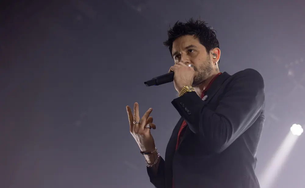 Man singing into a microphone on stage, wearing a suit jacket and jewelry.