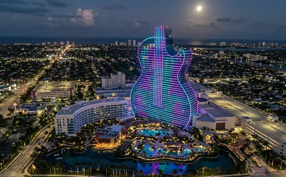 Night view of a guitar-shaped hotel lit with neon lights, surrounded by a resort and cityscape.