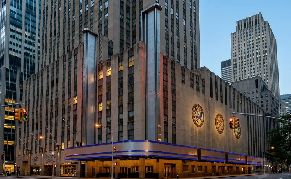 Art Deco buildings in NYC, featuring ornate mosaic medallions and illuminated blue trim, are seen at dusk.