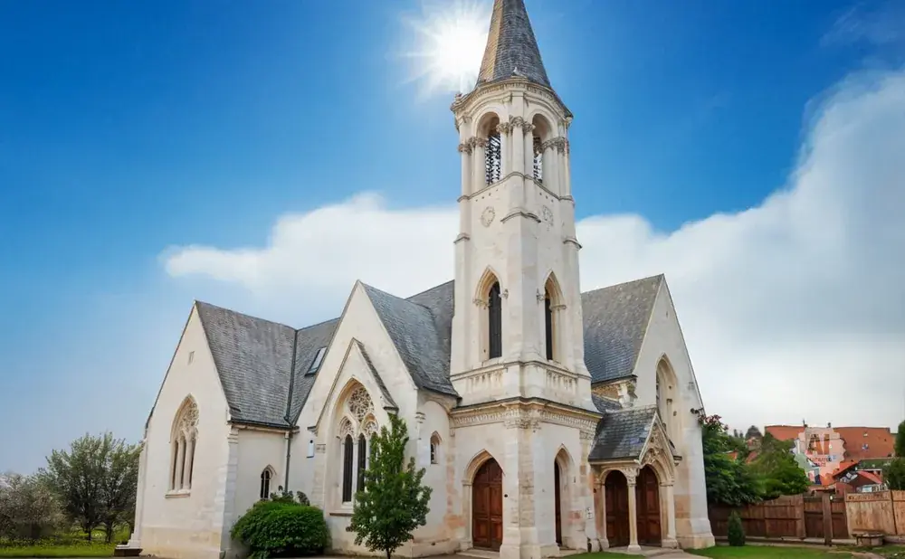 Exterior of a pale stone church with a tall steeple, under a bright blue sky with the sun shining.