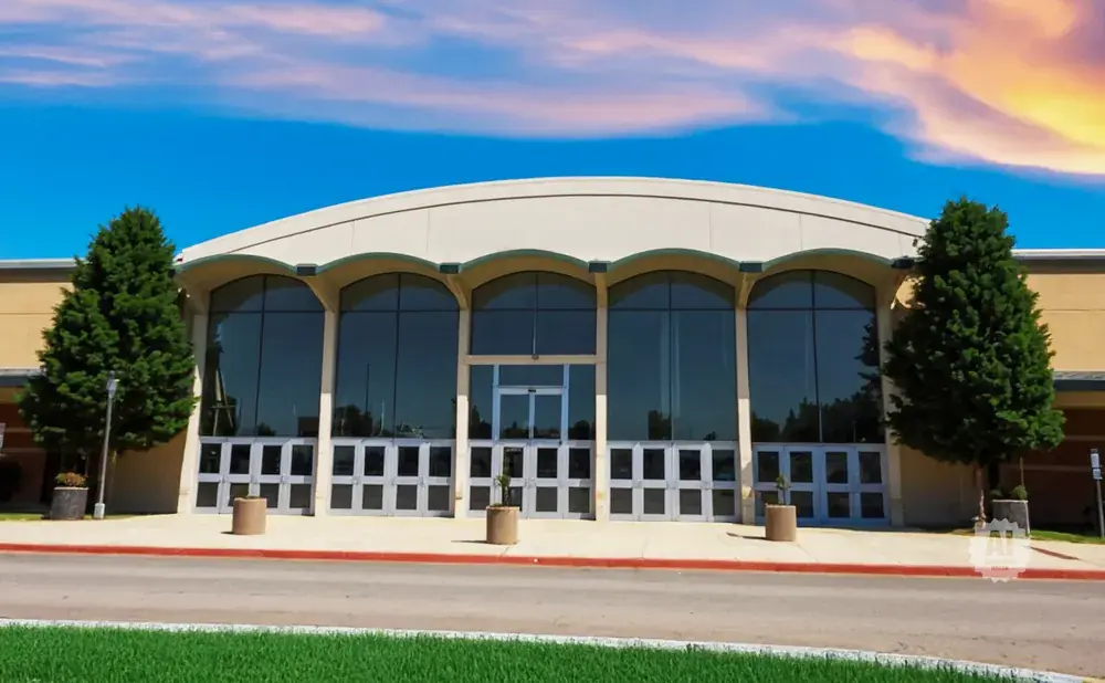 Modern building with large arched windows and glass doors, flanked by trees under a colorful sky.