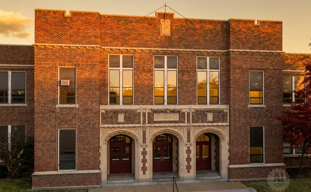 Exterior of an old brick school building with arched doorways and many windows reflecting the sunset.