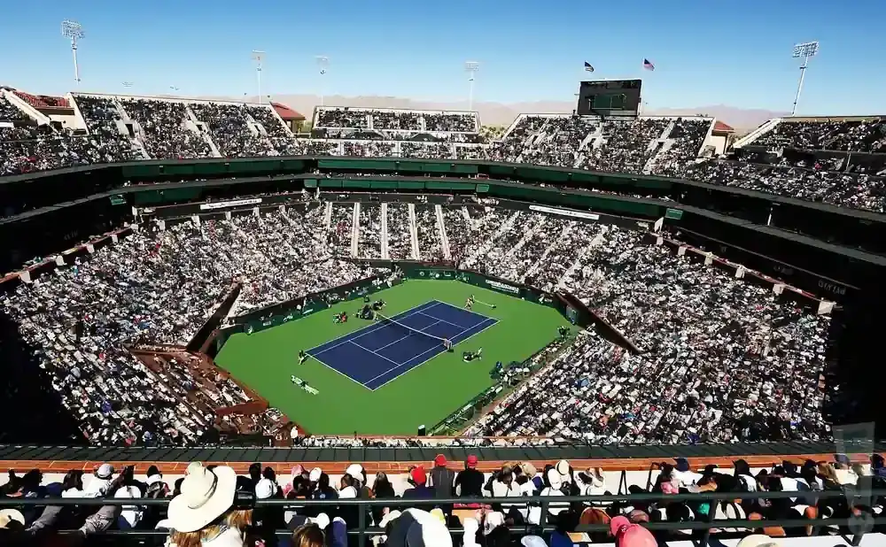 Aerial view of a packed tennis stadium with a match in progress on a bright, sunny day.