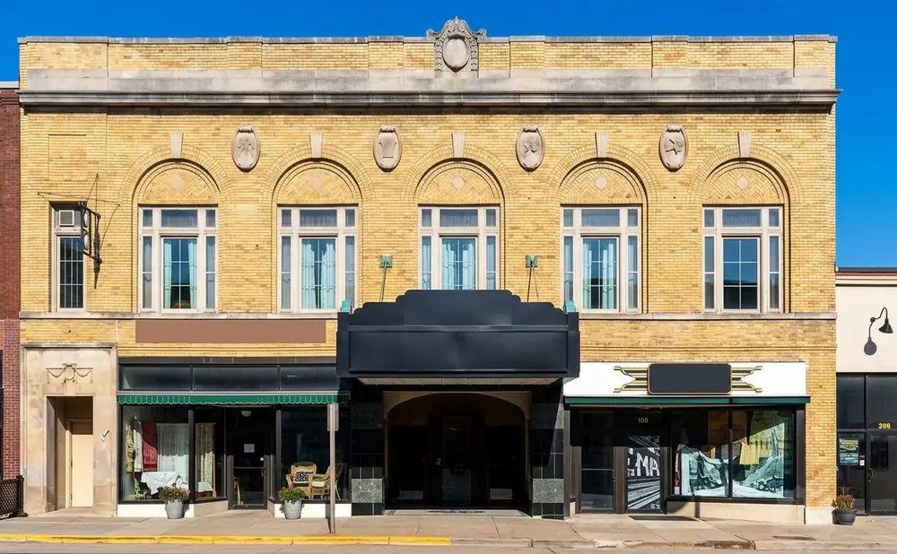 Yellow brick building with arched windows, a black marquee over an entrance, and storefronts below.