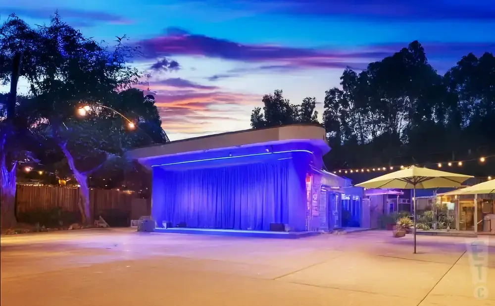 A stage with blue curtains is lit up at dusk, with trees and string lights in the foreground.