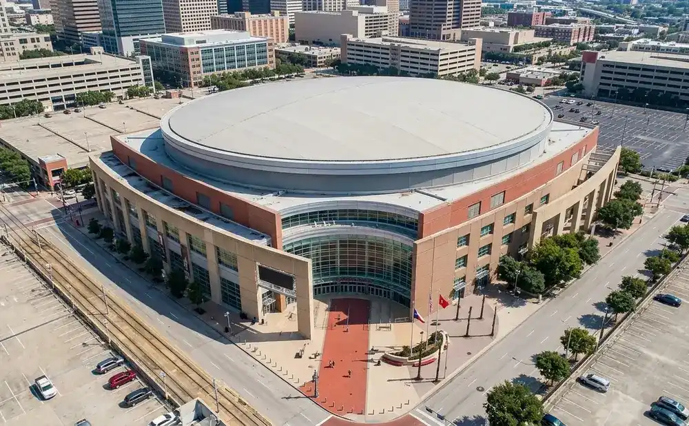 Aerial view of a large, circular arena with a gray roof and a beige and red exterior, surrounded by parking lots and city buildings.