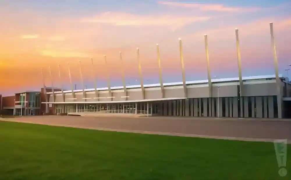 A modern building with a long glass facade stands under a colorful sunset sky, with a green lawn in the foreground.