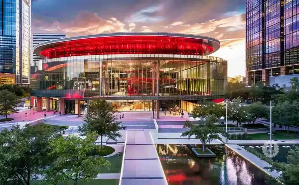 Modern glass building with a red-lit top level, surrounded by trees and a reflective pool.