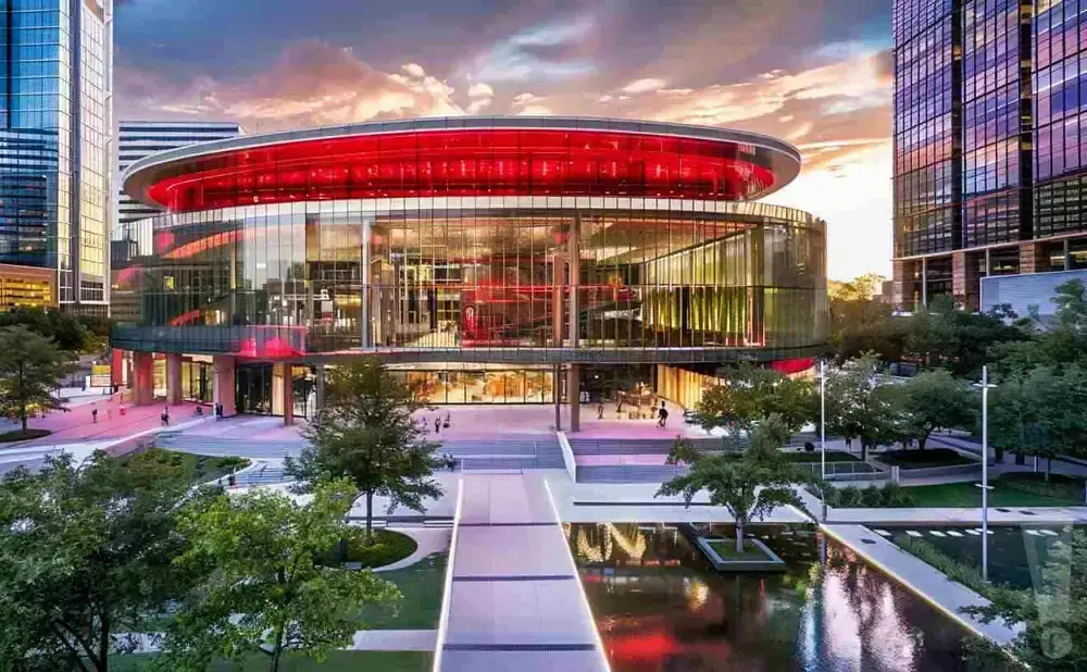 A modern, circular glass building with a red roof, surrounded by trees and reflecting pools, at sunset.