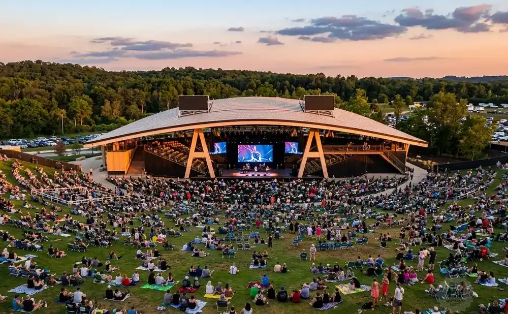 Outdoor concert with a packed lawn seating area and a large stage under a curved roof.