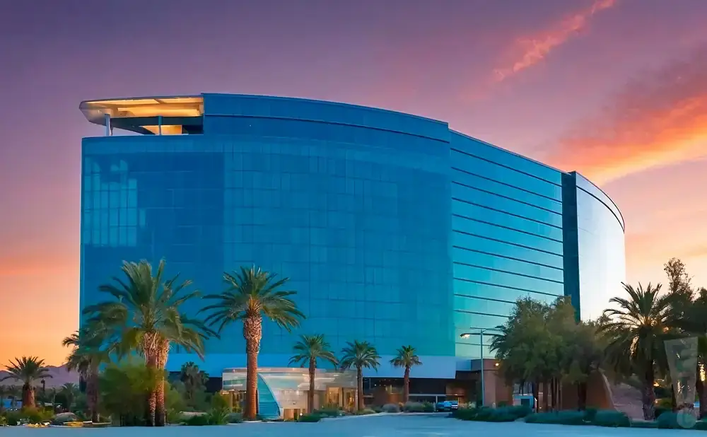 A modern, blue-glass hotel with palm trees in front against a vibrant sunset sky.