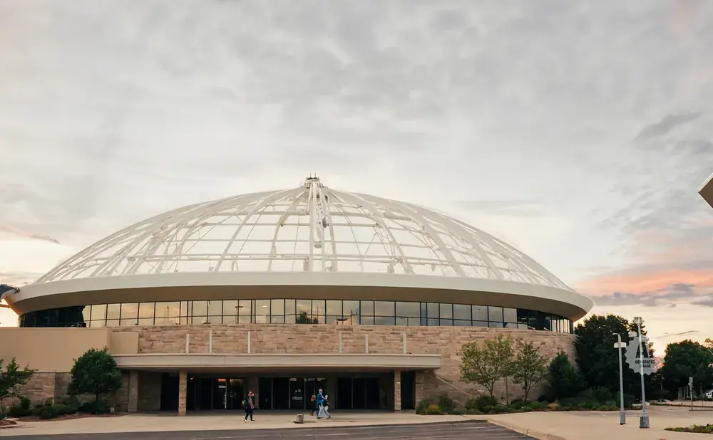 A modern domed building with a stone facade and large windows, under a cloudy sky at dusk.