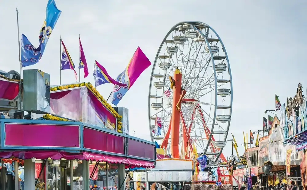 A Ferris wheel and carnival rides at a fair under a light sky, with colorful flags flying.