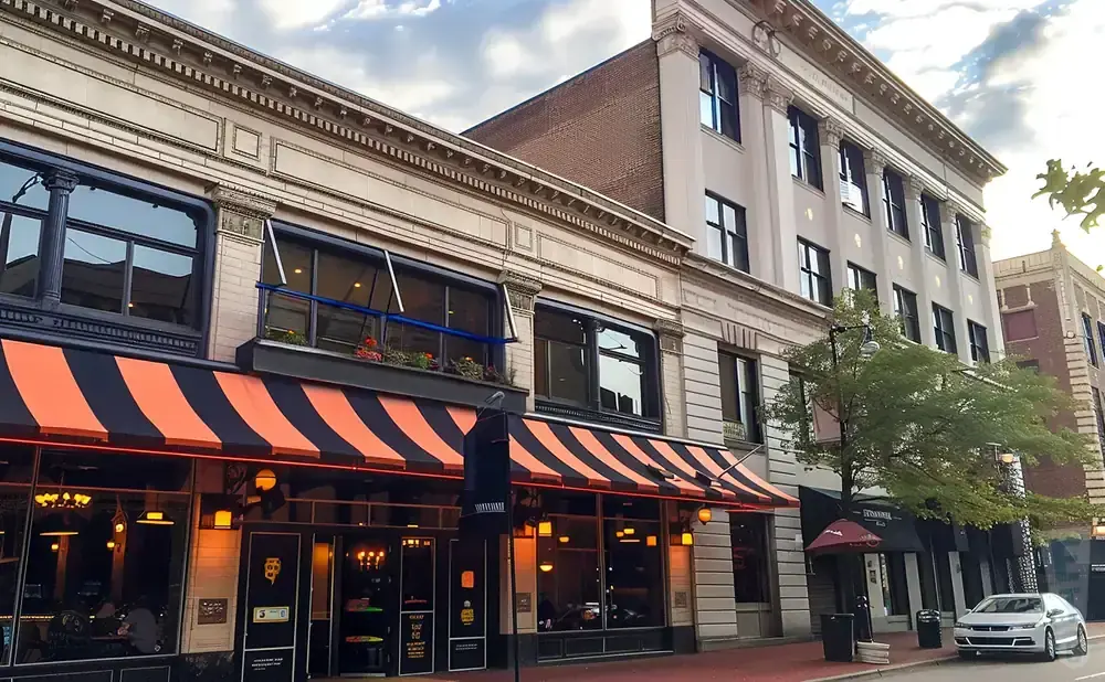 A row of historic buildings with orange and black awnings on a street. A silver car is parked on the side.