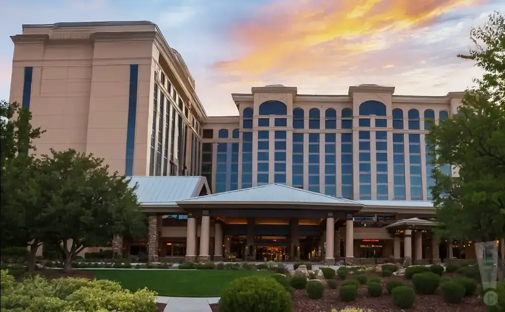 A large tan hotel with a blue-windowed facade and a covered entrance, framed by trees and a dramatic sunset sky.