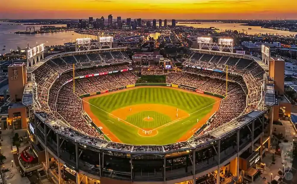 Aerial view of a baseball stadium at sunset with a city skyline and bay in the background.