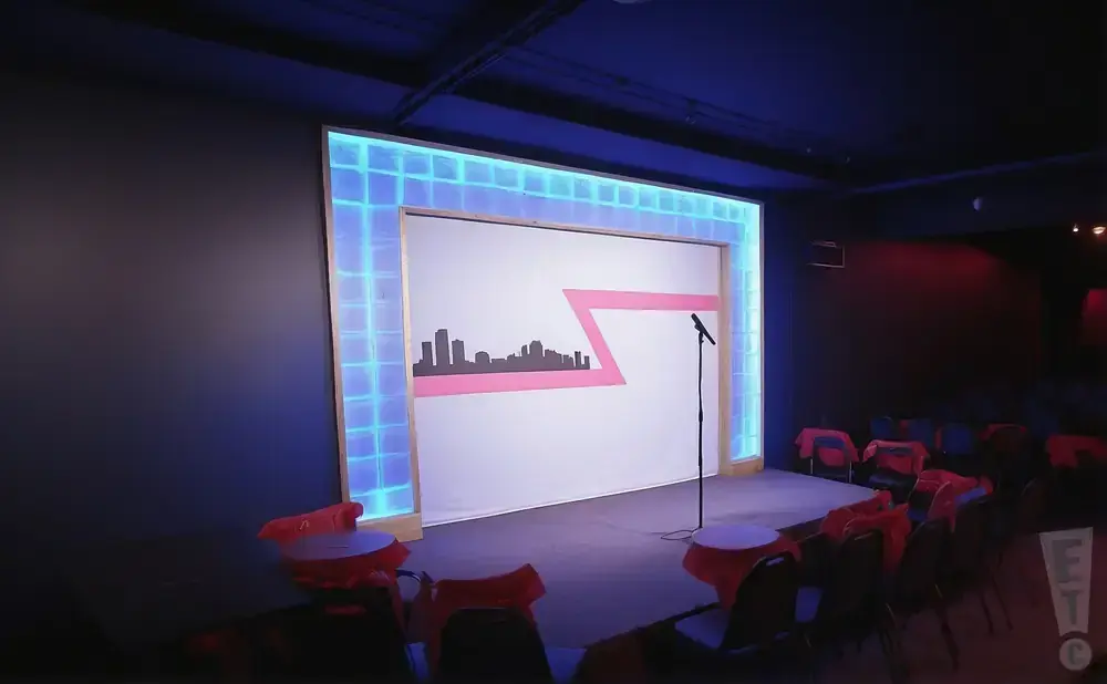 A comedy club stage with a cityscape backdrop, a microphone, and red-draped tables.