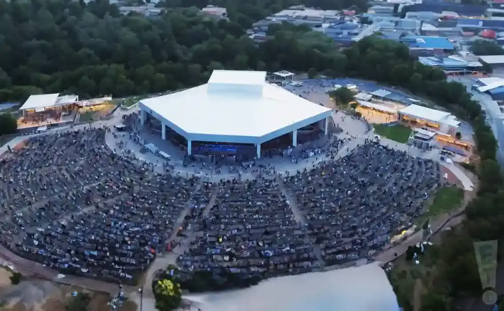 Aerial view of an outdoor amphitheater with a large white-roofed stage and a full audience in tiered seating.
