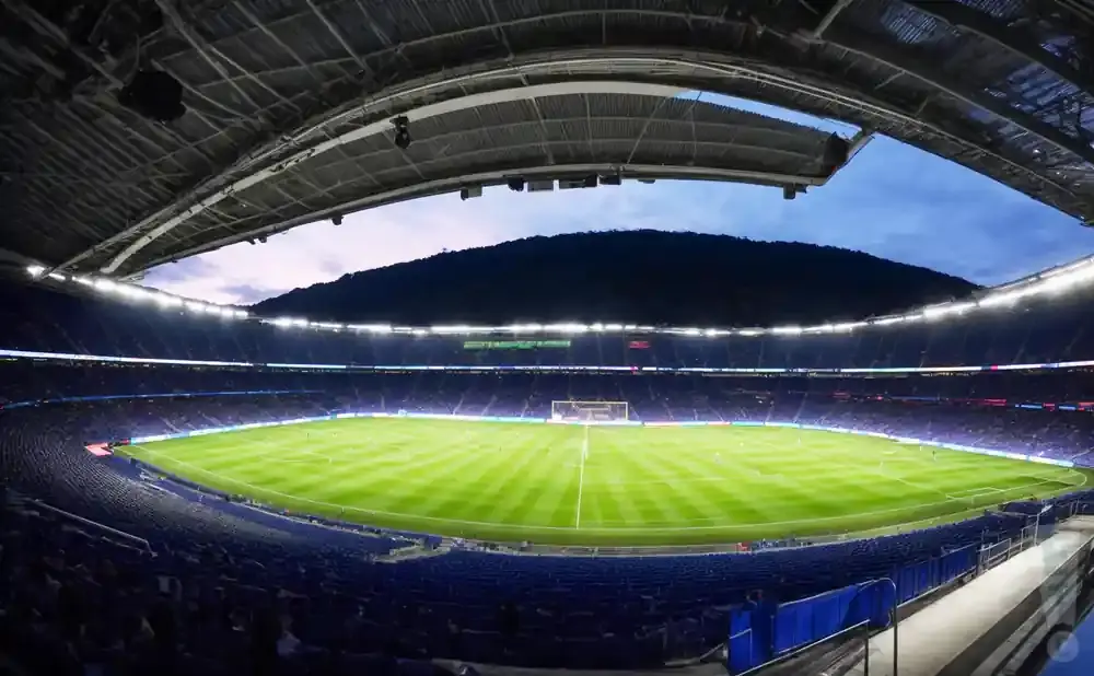 Wide angle view of a soccer stadium with a lit green field and blue seats, under a dusky sky.