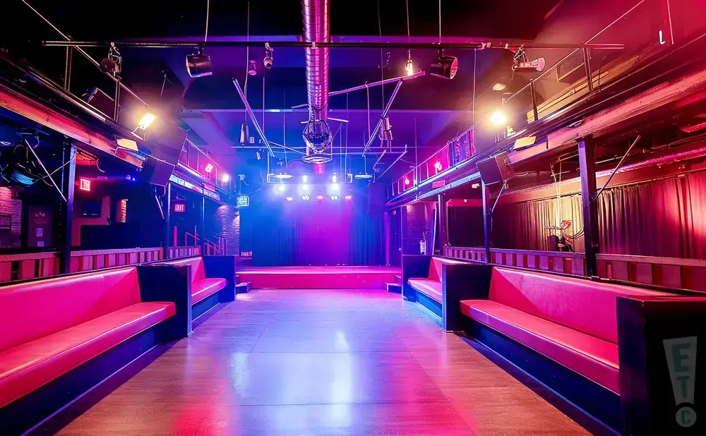 A dimly lit nightclub stage with a bright purple and blue spotlight, red seating, and a disco ball.