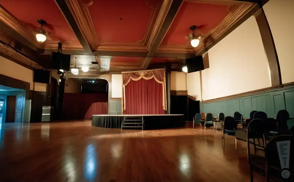 A wide view of an empty ballroom with a stage and red curtains, set up with chairs and screens.