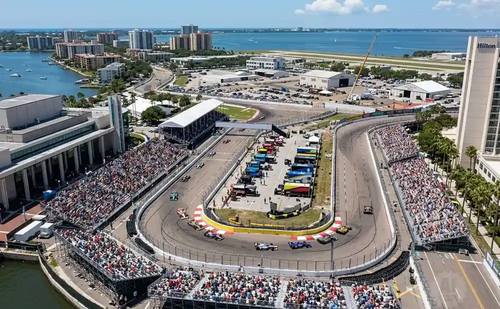 An aerial view of a race track with cars and spectators, set against a backdrop of city buildings and the ocean.