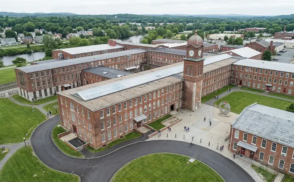 Aerial view of a large brick industrial complex with a clock tower, river, and surrounding trees.