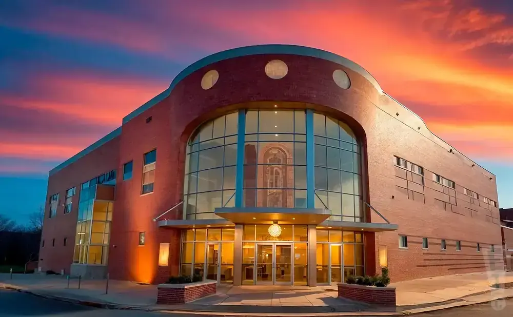 A modern brick building with a large glass entrance is lit up against a vibrant sunset sky.
