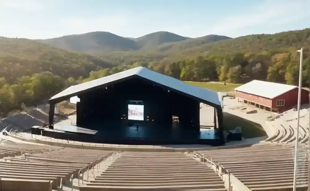 Outdoor amphitheater with a large stage, seating, and a red barn in the background, framed by rolling hills.
