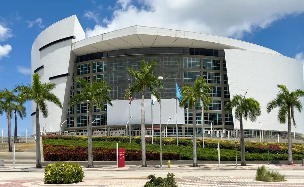 Modern white arena with glass facade, palm trees, and landscaped planters under a blue sky.
