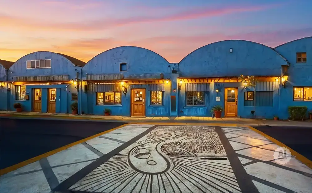 Blue buildings with arched roofs and lit windows under a sunset sky, with a mural on the pavement in front.