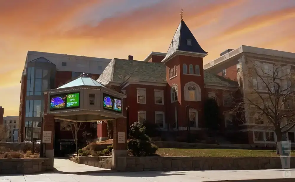 An old brick building with a tower and a modern glass addition is under a colorful sunset sky.