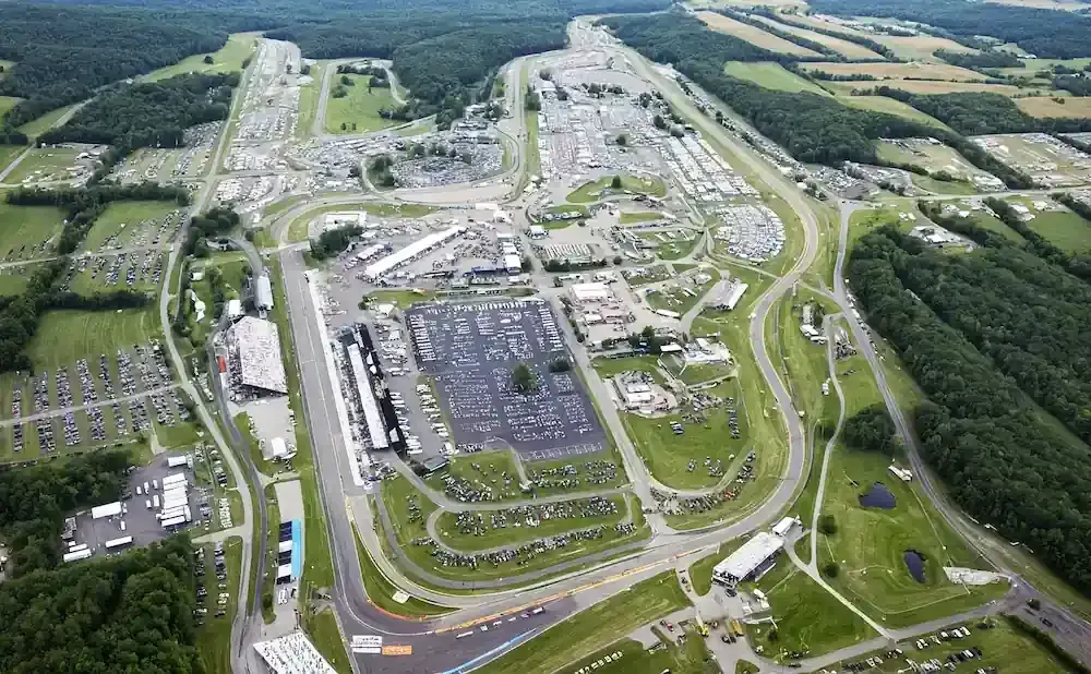 Aerial view of a large race track with many grandstands, parking lots full of cars, and surrounding forest.