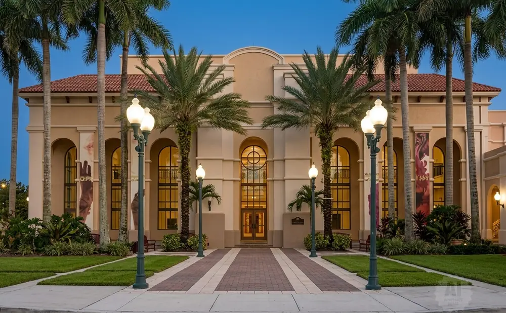 Exterior of a tan building with arched windows and red tile roof, flanked by palm trees and lit by ornate lampposts.