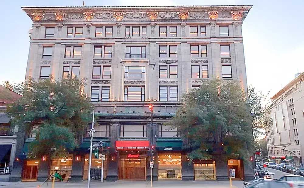 An ornate, multi-story stone building with many windows, framed by trees and a street scene with cars.