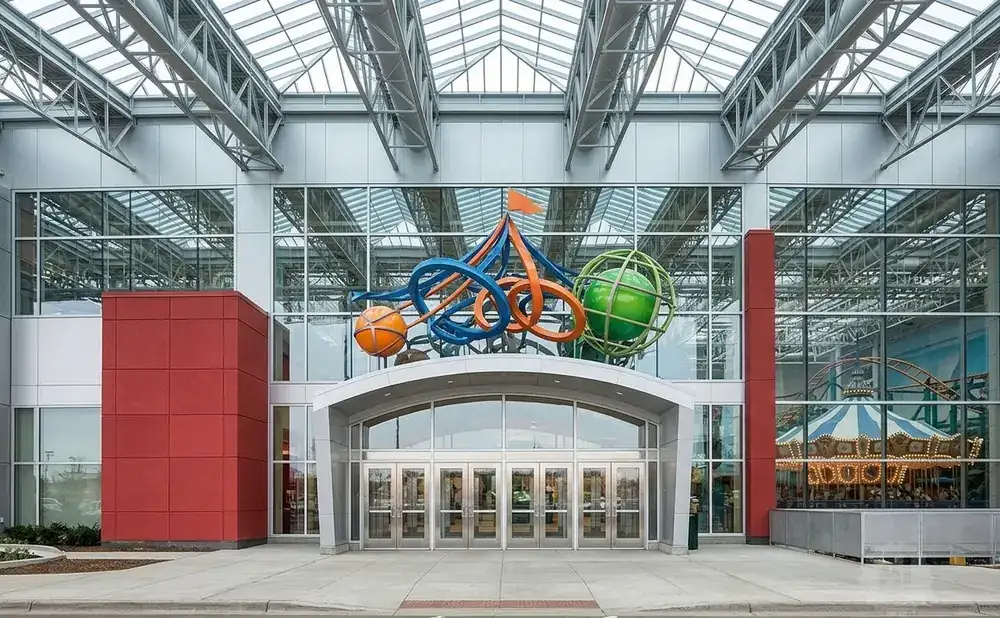 Modern building entrance with glass facade, abstract sculpture, and amusement park rides visible inside.