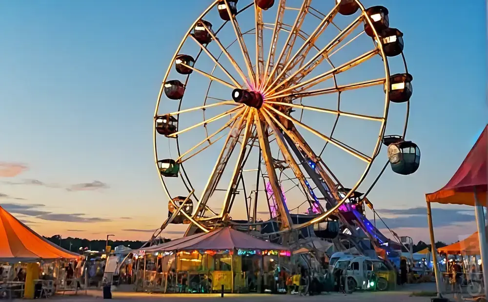 A Ferris wheel illuminated at dusk at a fairground, with tents and rides below.
