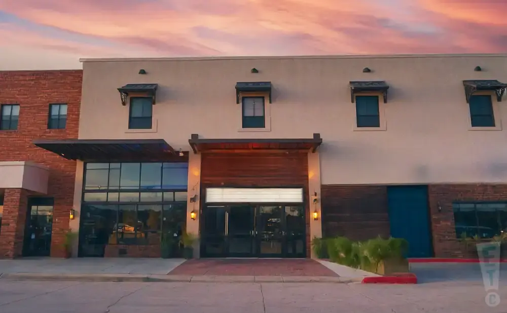 A modern building with brick and stucco exterior, large glass doors, and windows under a pink sunset sky.